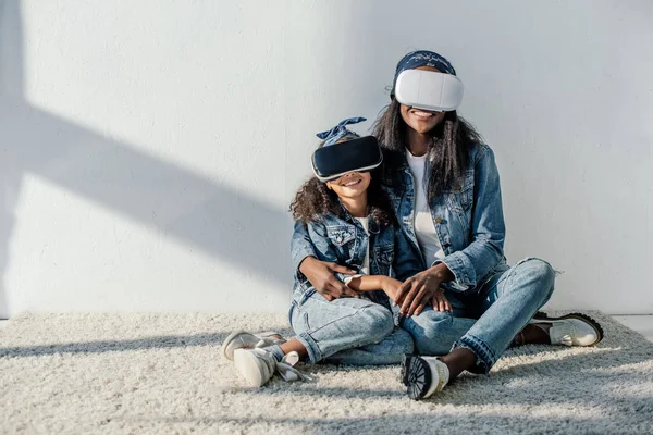 African american mother hugging daughter in similar clothing and vr headsets at home — Stock Photo