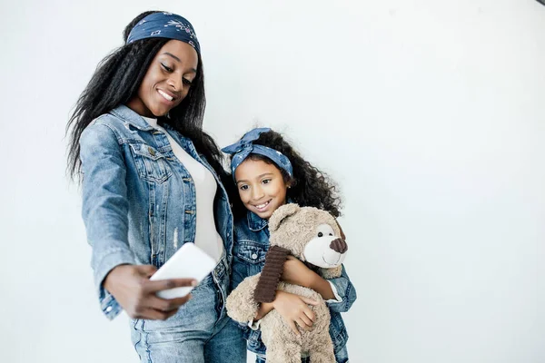 Smiling african american woman and daughter in similar clothing taking selfie on smartphone — Stock Photo