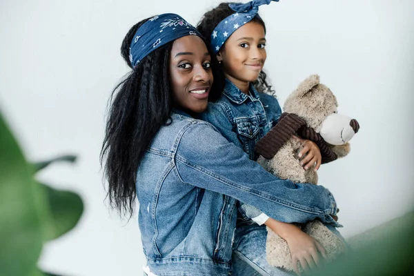 Portrait of cheerful african american woman hugging little daughter with teddy bear at home — Stock Photo