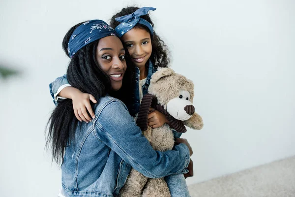 Portrait d'une fille afro-américaine souriante avec un ours en peluche embrassant sa mère à la maison — Photo de stock