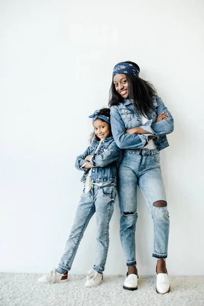 Sonrientes afroamericanos madre e hija con ropa similar posando en la pared en casa - foto de stock