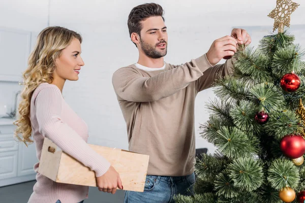 Paar schmückt Weihnachtsbaum zu Hause gemeinsam mit Kugeln, Freundin hält Holzkiste mit Spielzeug — Stockfoto