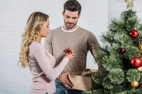Paar schmückt Weihnachtsbaum zu Hause gemeinsam mit Kugeln, Freund hält Holzkiste mit Spielzeug — Stockfoto