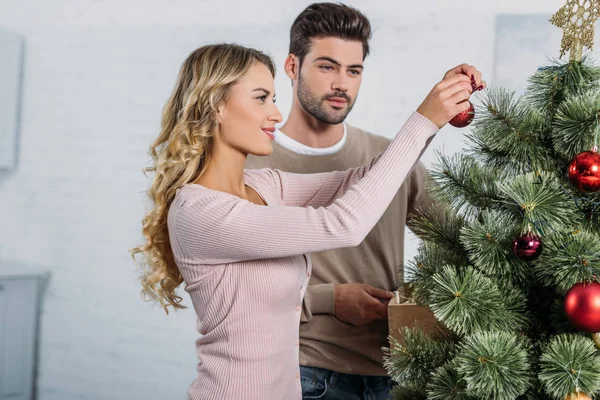 Girlfriend and boyfriend decorating christmas tree with baubles together at home — Stock Photo