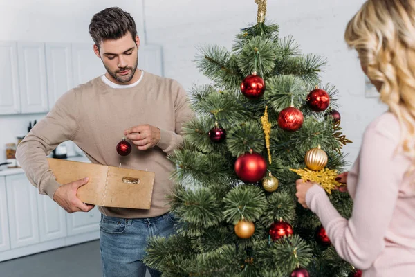 Handsome boyfriend holding wooden box with baubles and girlfriend decorating christmas tree with snowflake — Stock Photo