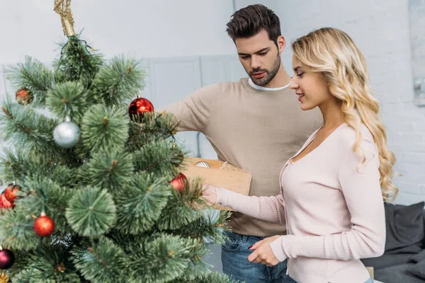 Boyfriend and girlfriend decorating christmas tree with baubles together at home — Stock Photo