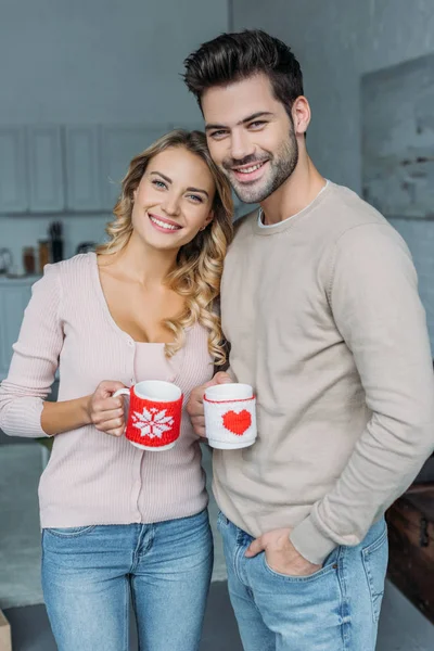 Sonriente pareja de pie con tazas de té en casa y mirando a la cámara, concepto de Navidad - foto de stock