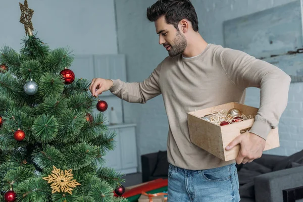 Schöner Mann schmückt Weihnachtsbaum mit Kugeln und hält Holzkiste zu Hause — Stockfoto