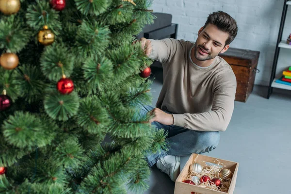 Homem bonito decorando árvore de Natal com bugigangas e sentado no chão em casa — Fotografia de Stock