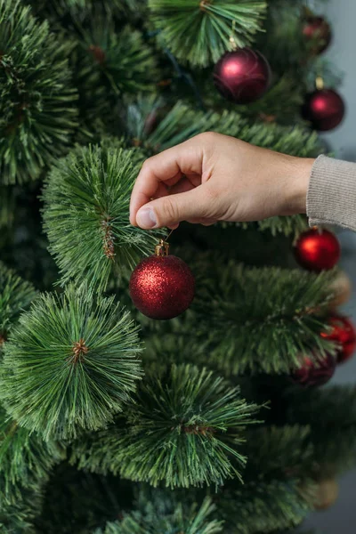 Imagen recortada de hombre decorando árbol de Navidad con bolas rojas en casa - foto de stock