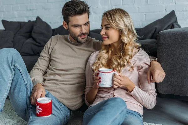 Smiling boyfriend and girlfriend holding cups of cappuccino and sitting on carpet in living room — Stock Photo