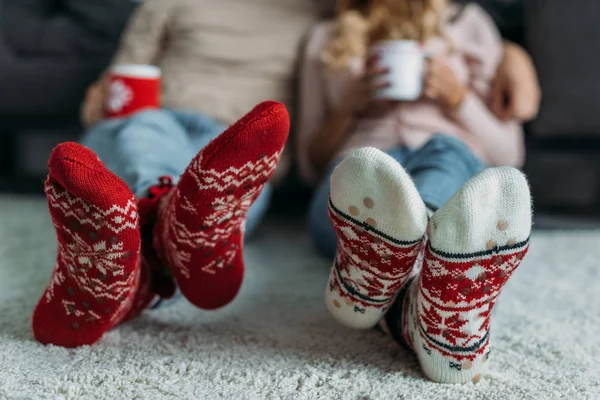 Imagen recortada de pareja sosteniendo tazas de capuchino con calcetines de Navidad en primer plano en casa - foto de stock