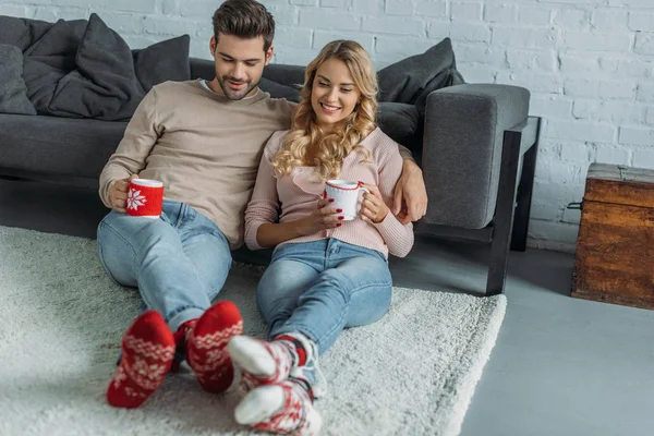 Smiling couple holding cups of cappuccino and sitting on carpet in living room — Stock Photo