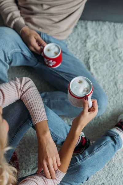 Обрезанное изображение пары, сидящей на ковре и держащей чашки капучино дома, рождественская концепция — стоковое фото
