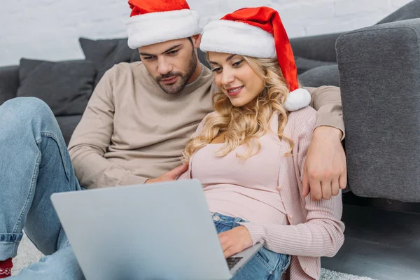 Novio y novia en sombreros de santa usando el ordenador portátil cerca del sofá en casa - foto de stock