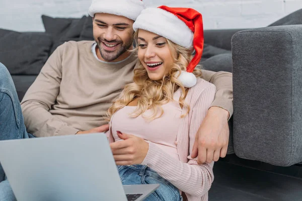 Casal animado em chapéus de santa usando laptop perto de sofá em casa — Fotografia de Stock