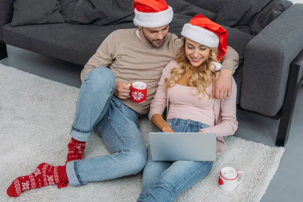 Blick auf ein glückliches Paar in Weihnachtsmannmützen, das auf dem Boden sitzt und zu Hause Laptop benutzt — Stockfoto