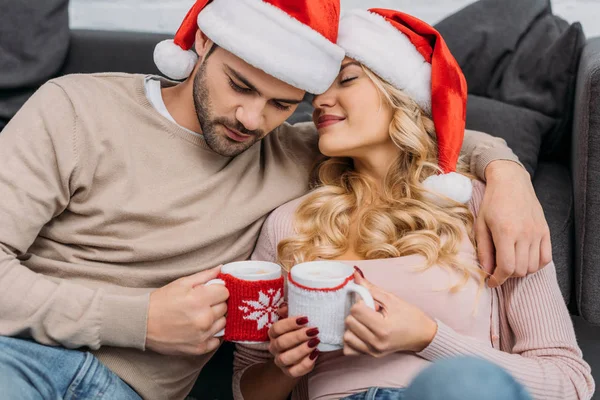 Liebevolles Paar in Weihnachtsmannmützen mit Cappuccino-Tassen zu Hause — Stockfoto