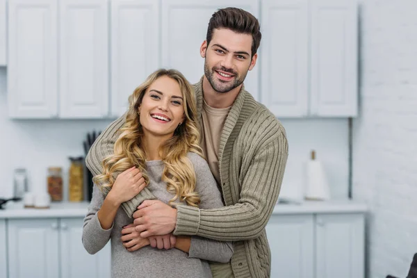 Sorrindo bonito namorado abraçando namorada atraente na cozinha e eles olhando para a câmera — Fotografia de Stock