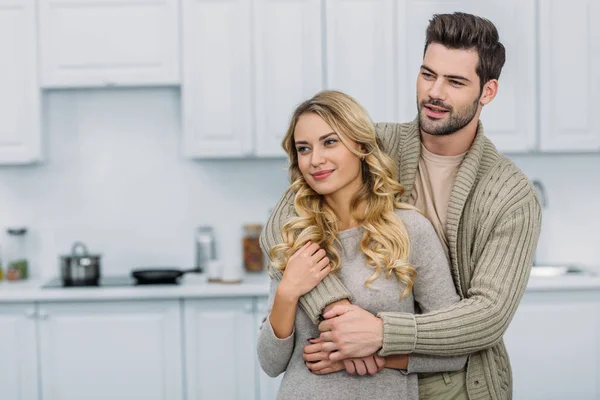 Handsome boyfriend hugging attractive girlfriend in kitchen and they looking away — Stock Photo