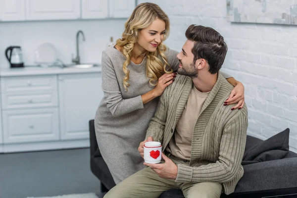 Girlfriend hugging boyfriend and he holding cup of coffee in kitchen — Stock Photo