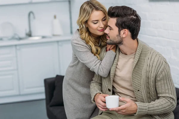 Girlfriend hugging boyfriend and he holding cup of tea in kitchen — Stock Photo