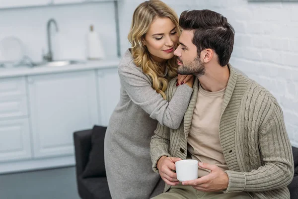 Petite amie étreignant petit ami et il tenant une tasse de thé dans la cuisine — Stock Photo