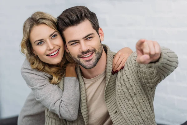 Petit ami souriant pointant sur quelque chose à petite amie sur le canapé dans le salon — Photo de stock