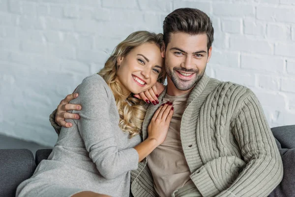 Portrait of smiling couple hugging and looking at camera on sofa in living room — Stock Photo