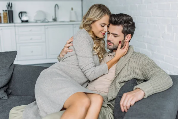 Beautiful young woman embracing her boyfriend while sitting on him on couch at home — Stock Photo