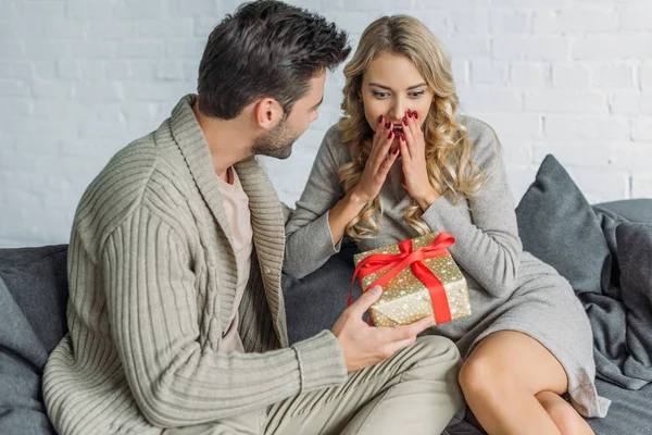 Handsome young man presenting gift for surprised girlfriend at home — Stock Photo