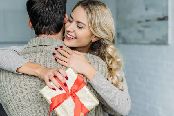 Attraktive junge Frau mit Geschenkbox umarmt ihren Freund zu Hause — Stockfoto