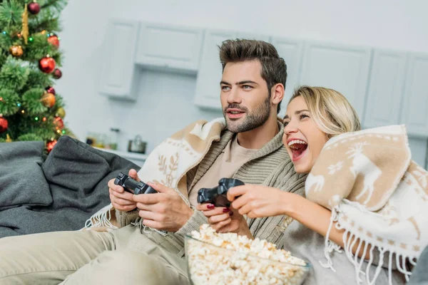 Feliz pareja joven con palomitas de maíz jugando videojuegos juntos en el sofá en casa - foto de stock