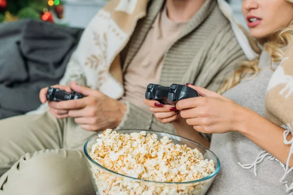 Recortado disparo de pareja con palomitas de maíz jugando videojuegos juntos en el sofá en casa - foto de stock