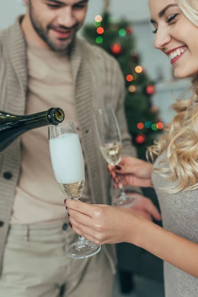 Hermosa pareja joven vertiendo champán en las copas delante del árbol de Navidad en casa — Stock Photo