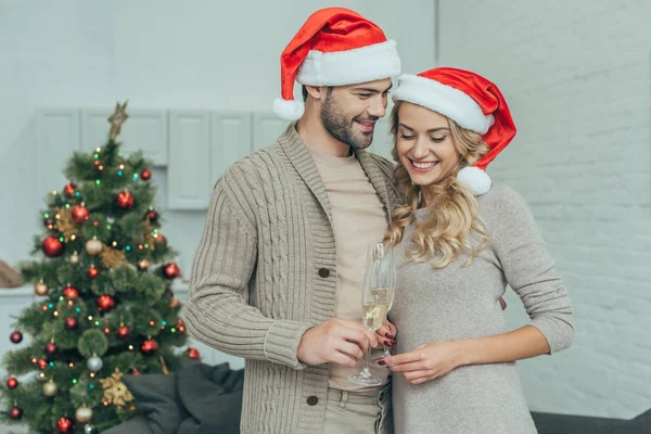 Heureux jeune couple avec des verres de champagne devant l'arbre de Noël à la maison — Photo de stock