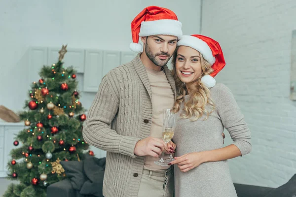 Happy young couple with champagne glasses looking at camera in front of christmas tree at home — Stock Photo