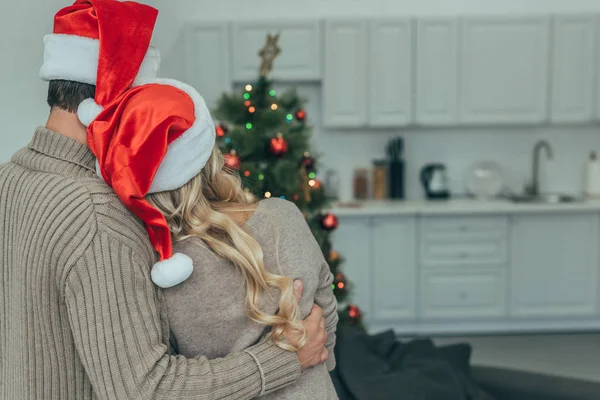 Rückansicht eines Pärchens mit Weihnachtsmützen, das zu Hause den Weihnachtsbaum betrachtet — Stockfoto