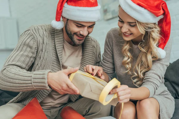 Творческая молодая пара в шляпах Санта-Клауса украшают рождественский подарок вместе дома — стоковое фото