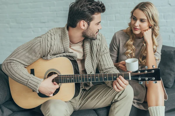 Atractivo joven tocando la guitarra para su novia en casa mientras ella está sentada en el sofá con café - foto de stock