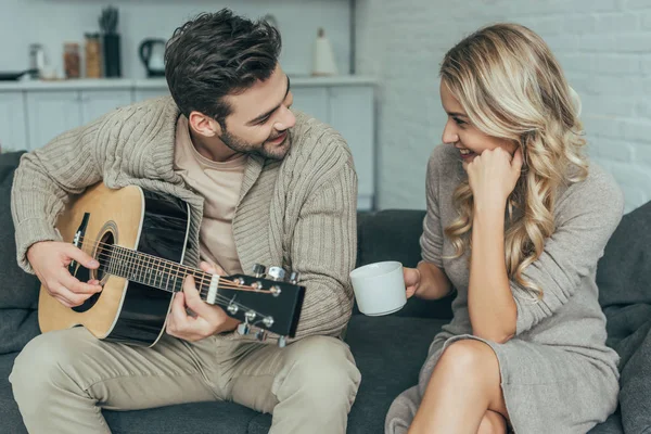 Счастливый молодой человек играет на гитаре для подруги дома, пока она сидит на диване с кофе — стоковое фото