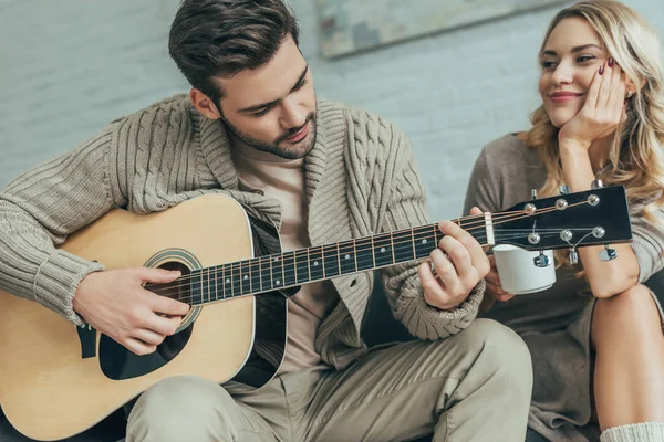 Jovem bonito tocando guitarra para a namorada em casa, enquanto ela está sentada no sofá com café — Fotografia de Stock