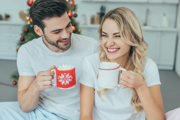 Смеющаяся молодая пара с чашками какао, сидящая в постели дома на Рождество — стоковое фото