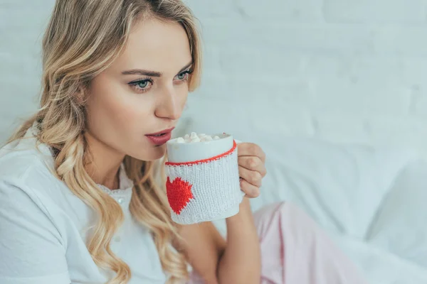 Beautiful young woman drinking cocoa with marshmallow in bed at home — Stock Photo
