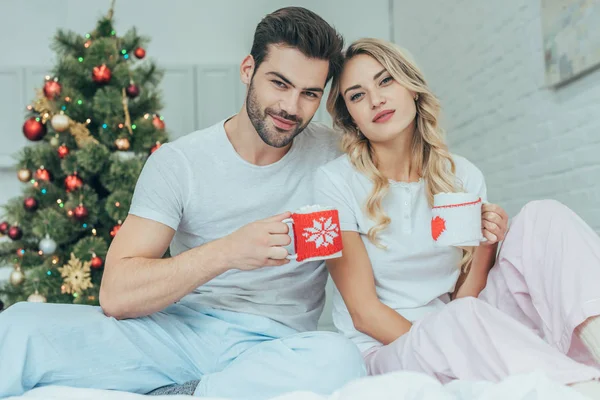 Beautiful young couple with cups of cocoa sitting in bed at home on christmas and looking at camera — Stock Photo