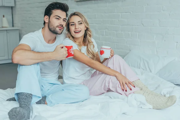 Hermosa pareja joven con tazas de cacao sentado en la cama en casa - foto de stock