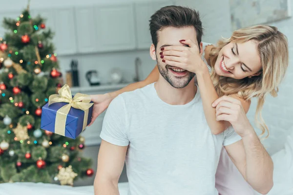 Hermosa joven mujer presentando regalo sorpresa de Navidad a novio en la cama en casa - foto de stock