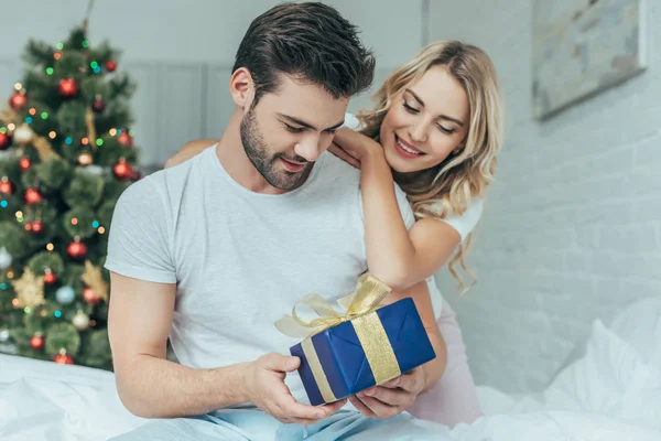 Hermosa joven mujer presentando regalo de Navidad a novio en la cama en casa - foto de stock