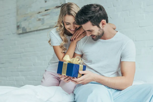 Schöne junge Frau überreicht Geschenk an Freund zu Hause im Bett — Stockfoto