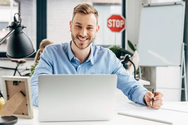 Guapo sonriente joven hombre de negocios utilizando el ordenador portátil y tomando notas en el lugar de trabajo - foto de stock